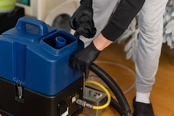 A person in gray pants and black gloves cleans a carpet using a blue machine in a home. A cloth is being used to assist in the cleaning process. The scene is well lit with natural light.